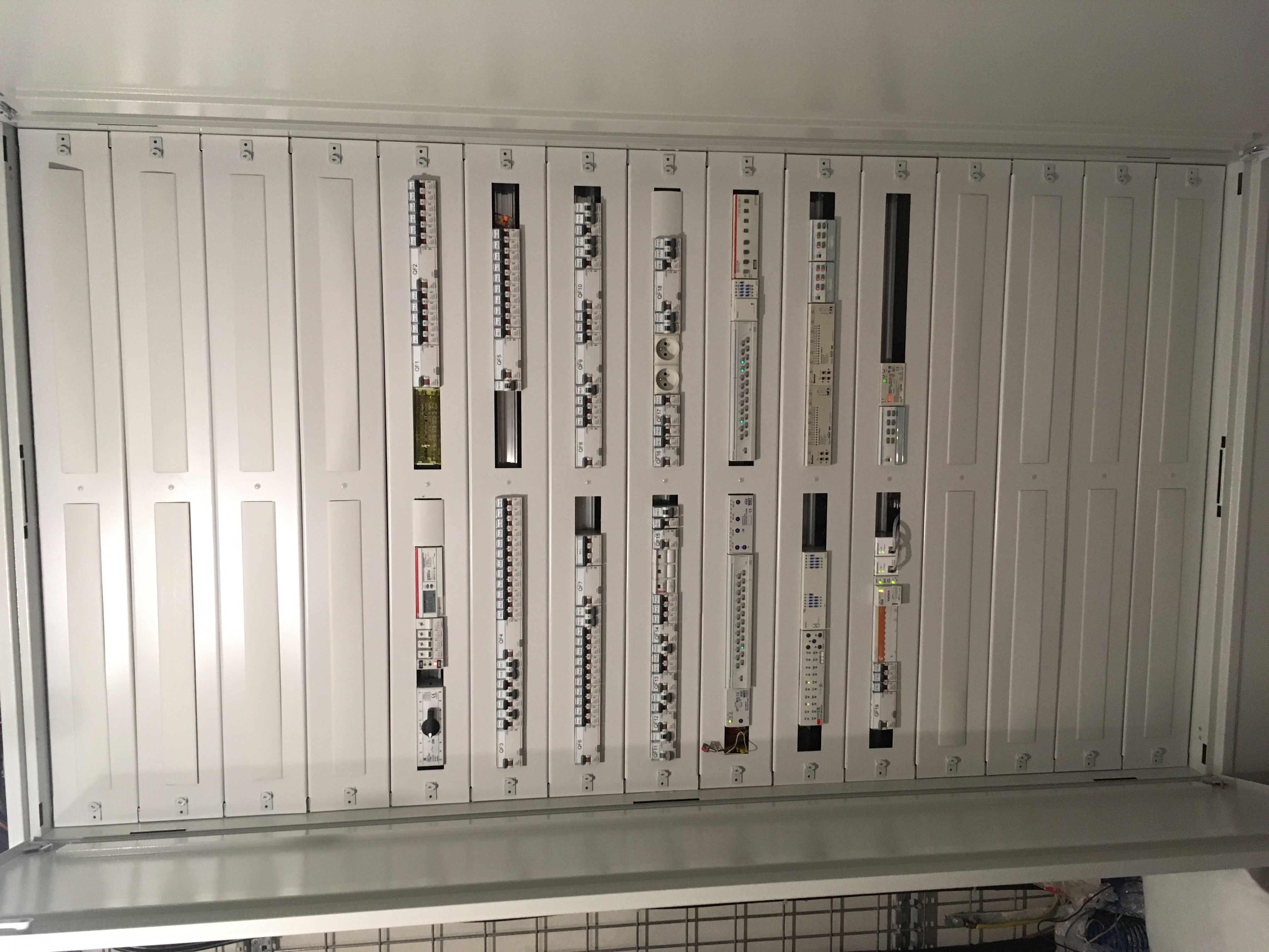 Otwarta, biała skrzynka rozdzielcza z zamontowanymi modułowymi urządzeniami elektrycznymi, widoczne bezpieczniki, wyłączniki różnicowoprądowe oraz inne komponenty z oznaczeniami QF1, QF2, QF3, QF4,...