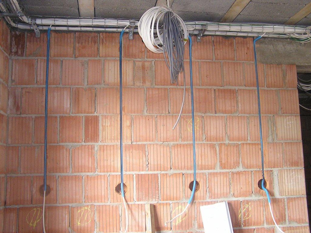 Surowa ściana z czerwonej cegły z pionowymi niebieskimi rurkami instalacyjnymi wpuszczonymi w otwory, kable poprowadzone pod sufitem, widoczne etapy prac instalacyjnych.
