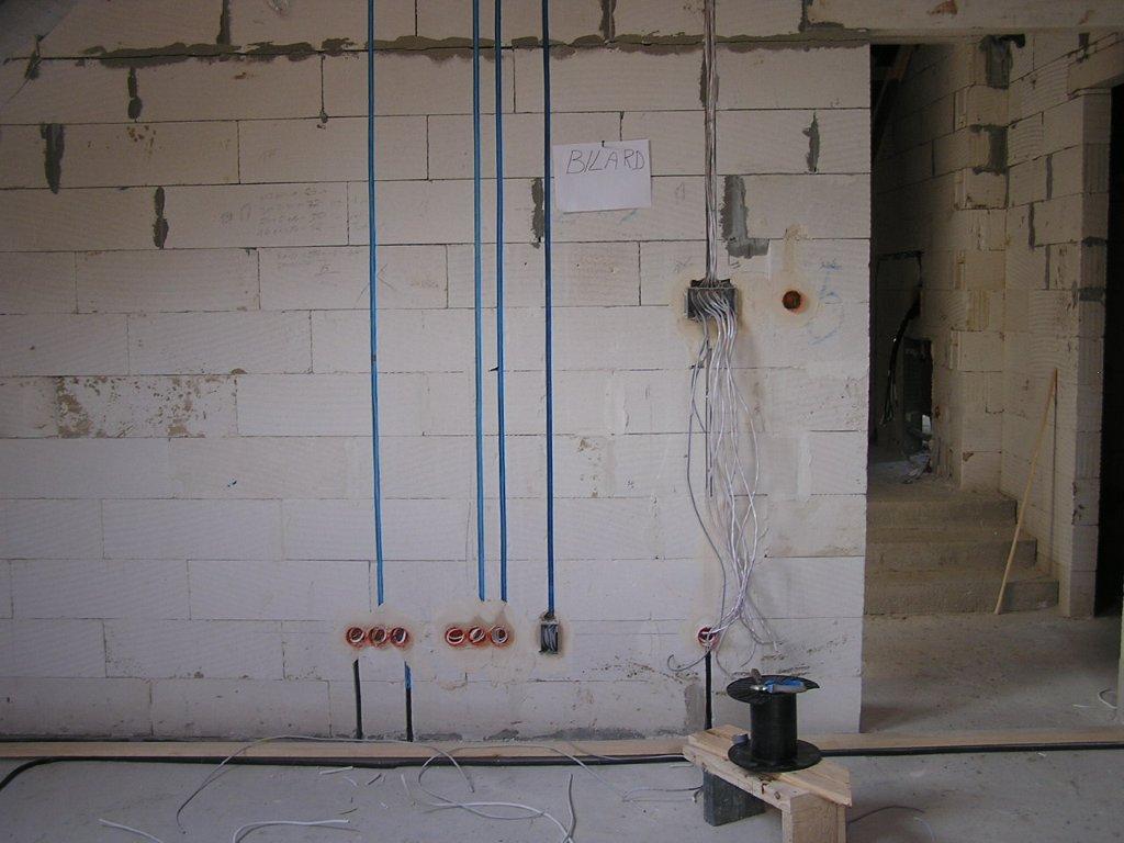 Surowa ściana z betonu komórkowego w trakcie prac elektrycznych: pionowe, niebieskie rurki instalacyjne, puszki podtynkowe i zwisające przewody. Na ścianie kartka z napisem 'BILARD'.