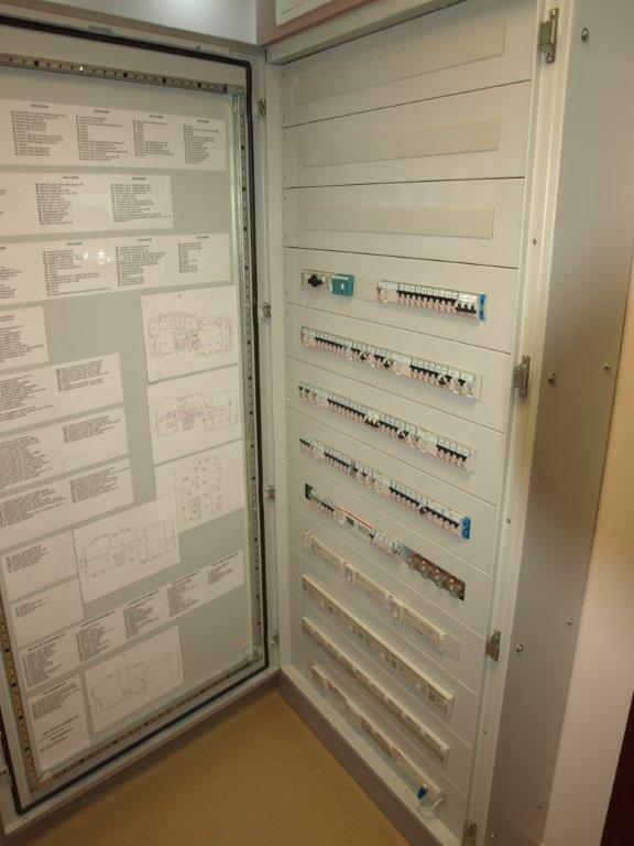 Otwarta szafa elektryczna z widocznymi bezpiecznikami, wyłącznikami i schematami instalacji na wewnętrznej stronie drzwi.