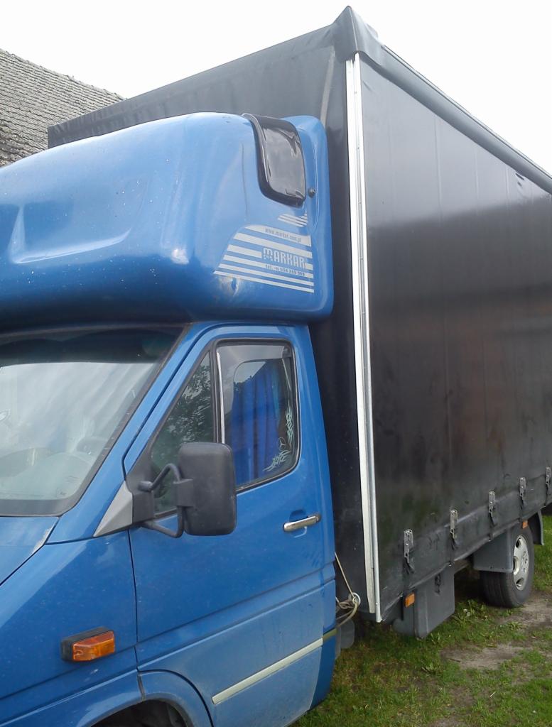 Niebieski samochód dostawczy z czarną plandeką, widoczne logo firmy na nadbudówce kabiny oraz fragment zielonego trawnika.