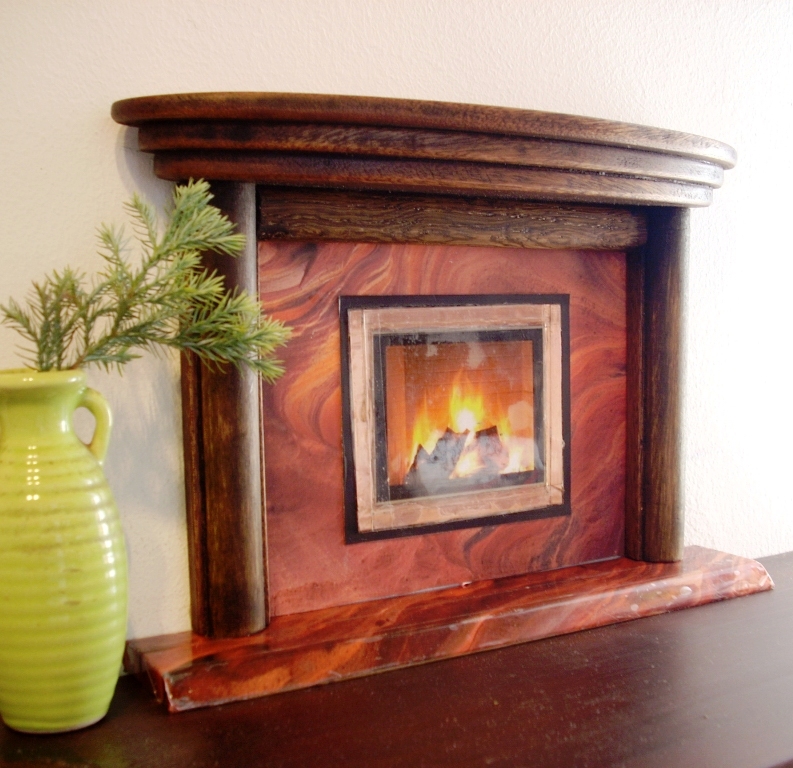 Dekoracyjny, drewniany portal kominkowy w miniaturze z płomieniami ognia za szybą, obok w wazonie gałązka świerku.