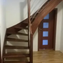 Kompaktowe, drewniane schody z metalową balustradą w jasnym wnętrzu. Widoczne drzwi z przeszkleniami i fragment wzorzystego dywanu.