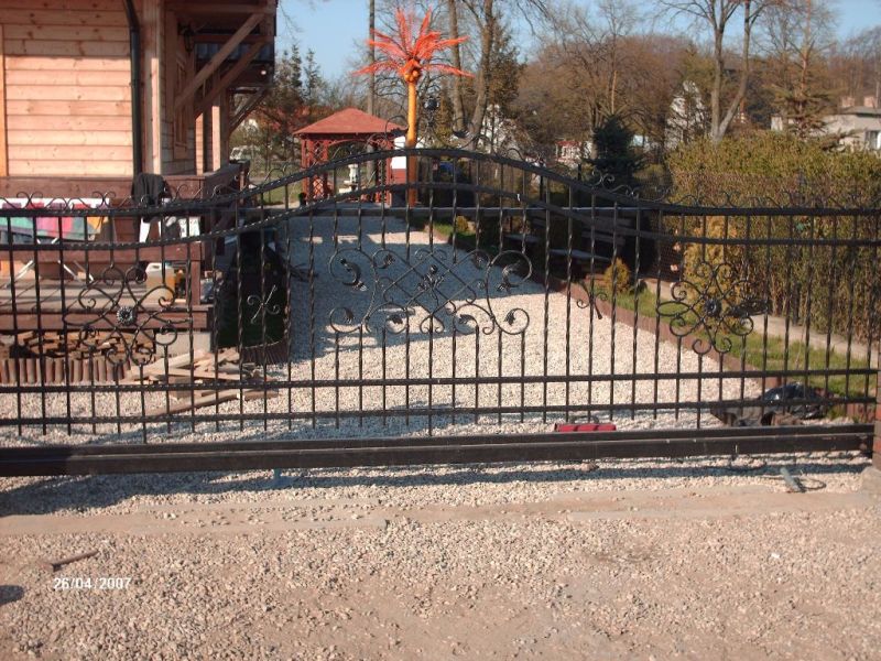 Czarna, metalowa brama przesuwna z ozdobnymi elementami kowalstwa artystycznego, prowadząca do posesji z drewnianym domem i altaną w tle.