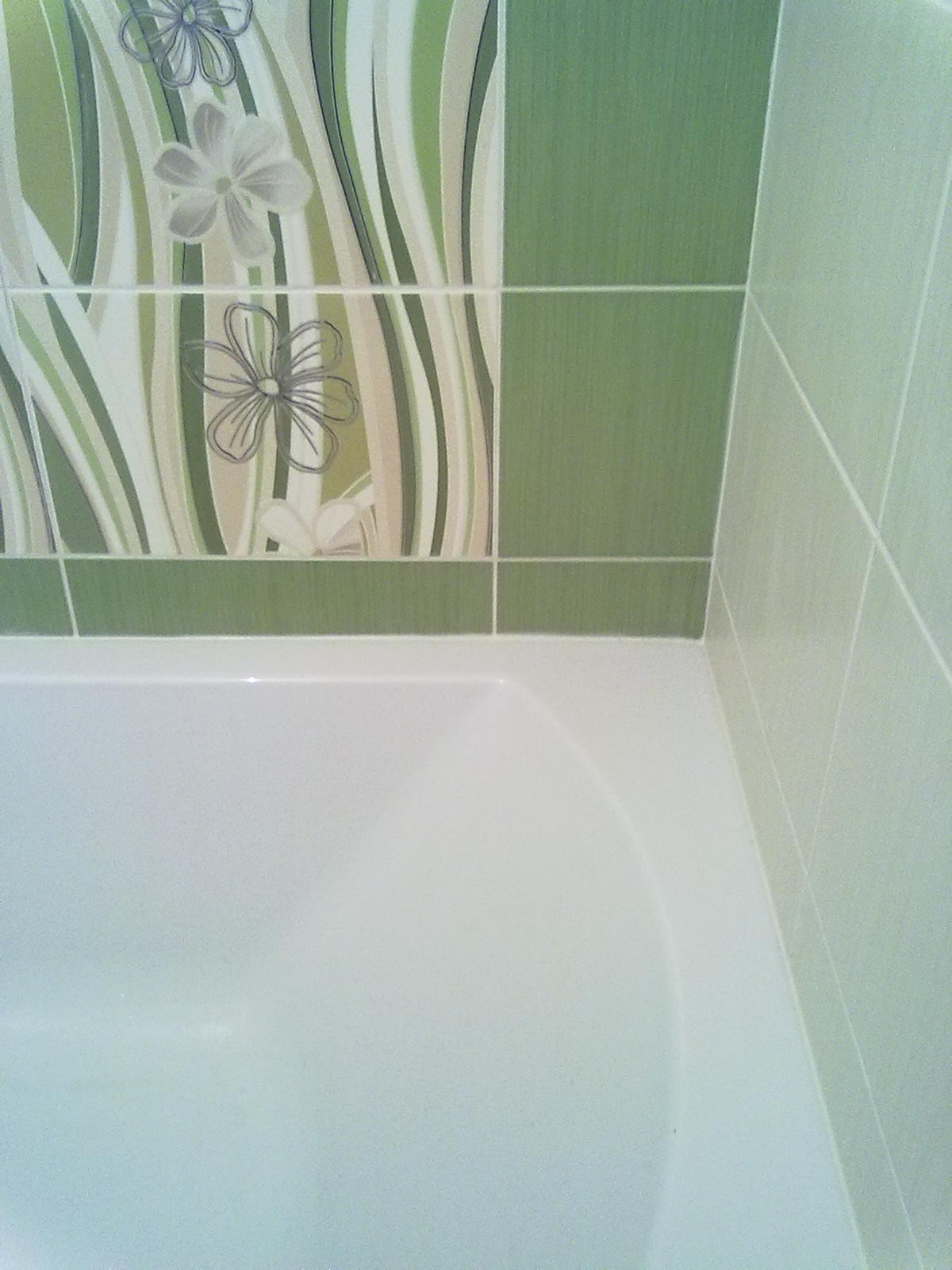 Fragment wykończonej łazienki: biała wanna z zielonymi płytkami z motywem kwiatowym na ścianie.