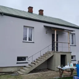 Dwupiętrowy dom z szarą elewacją po nałożeniu gładzi, widoczne schody wejściowe i przygotowane materiały budowlane na stole przed domem.