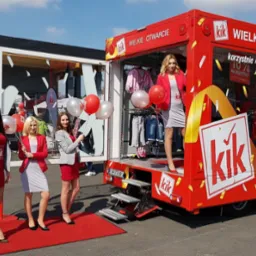 Czerwony, mobilny punkt sprzedaży detalicznej KiK podczas wielkiego otwarcia, z czterema młodymi kobietami w strojach promocyjnych trzymającymi czerwono-białe balony z logo KiK, stojącymi...