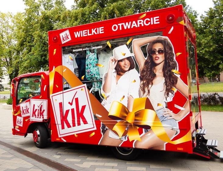 Czerwona ciężarówka z grafiką reklamową sklepu odzieżowego KiK, prezentująca hasło 'Wielkie Otwarcie', ubrania w witrynie i wizerunki dwóch modelek z kokardą, zaparkowana na chodniku.