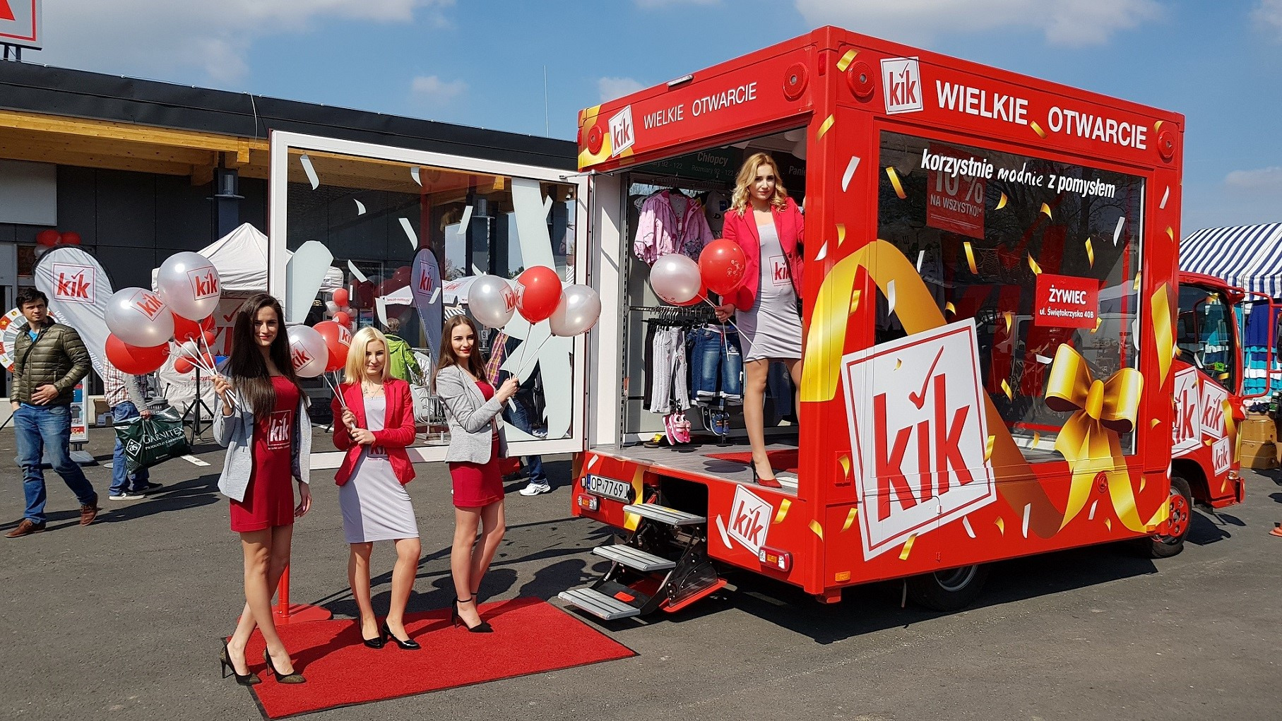 Czerwony, mobilny punkt sprzedaży detalicznej KiK podczas wielkiego otwarcia, z czterema młodymi kobietami w strojach promocyjnych trzymającymi czerwono-białe balony z logo KiK, stojącymi...