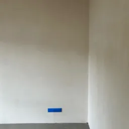 Wnętrze po szpachlowaniu: gładkie, jasne ściany w odcieniach beżu i szarości, z niebieską taśmą zabezpieczającą przy podłodze. Minimalistyczny kadr na r&oacute;g pomieszczenia.