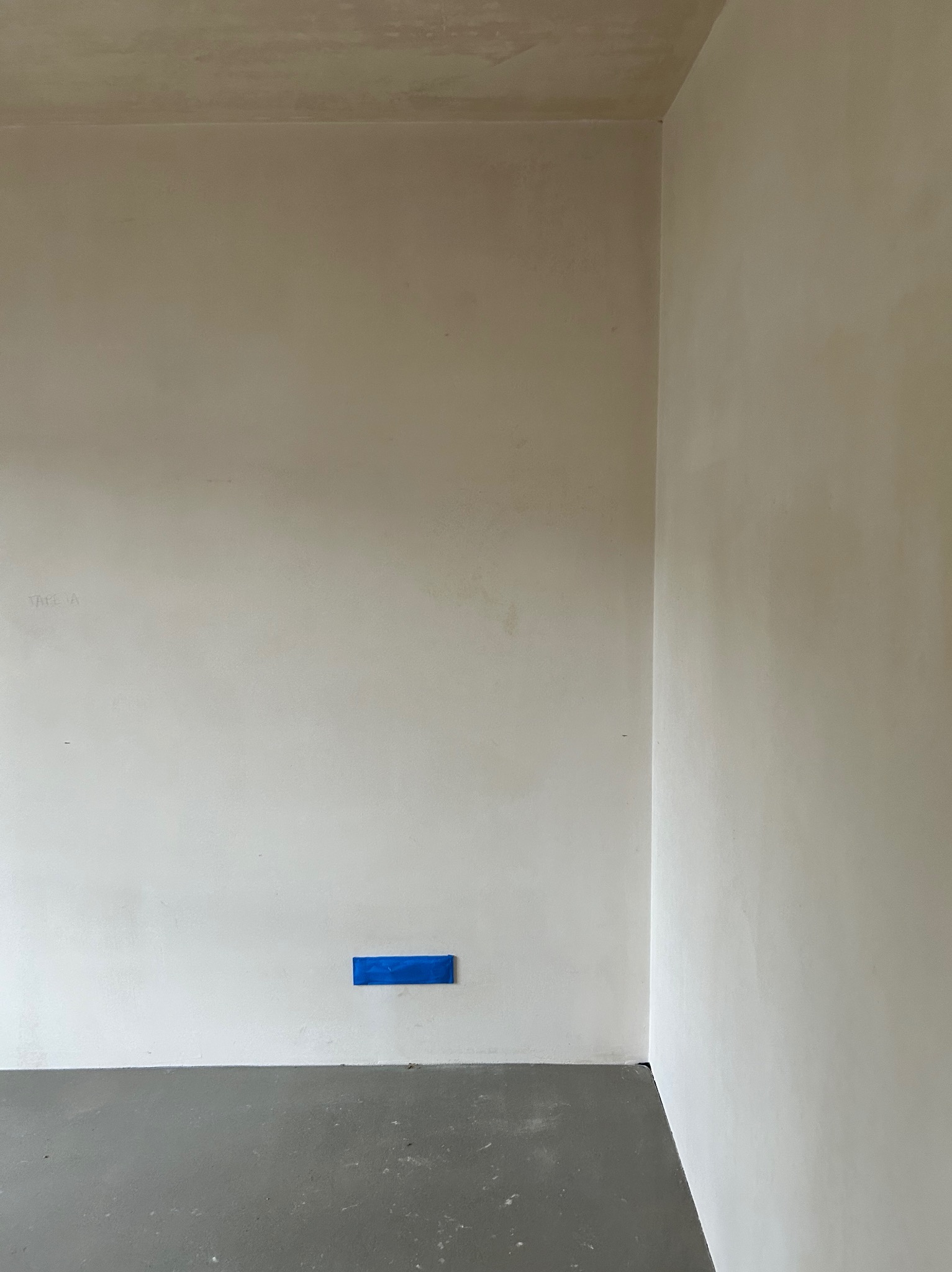 Wnętrze po szpachlowaniu: gładkie, jasne ściany w odcieniach beżu i szarości, z niebieską taśmą zabezpieczającą przy podłodze. Minimalistyczny kadr na róg pomieszczenia.