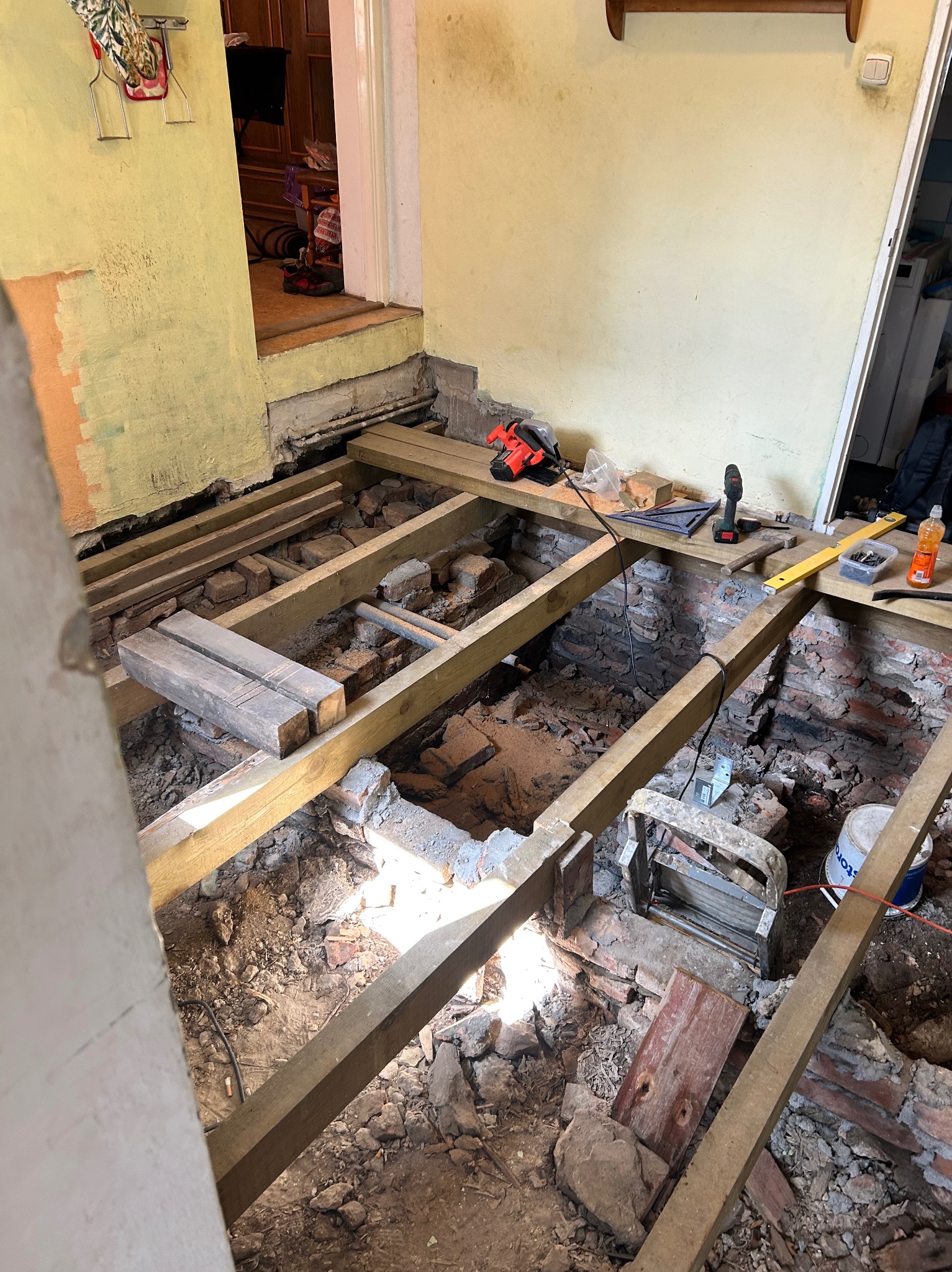 Drewniany szkielet podłogi w trakcie montażu, widoczne narzędzia i fragmenty cegieł. Ujęcie z góry, surowy stan wnętrza przed wykończeniem.