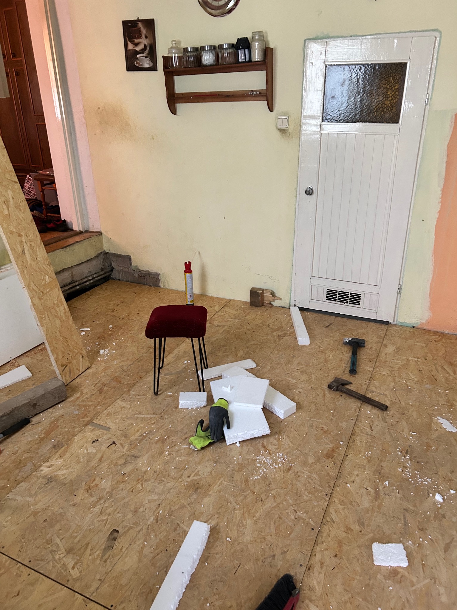 Układanie izolacji pod panele na podłodze z płyty OSB. Widoczne narzędzia, fragmenty styropianu i czerwony stołek. Remont wnętrza w trakcie prac.