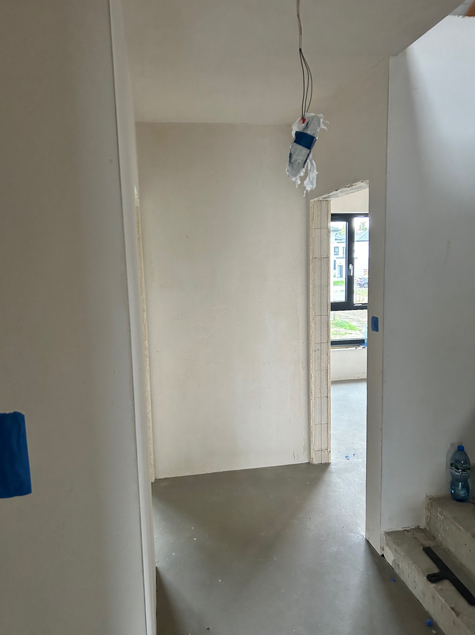 Wnętrze w trakcie wykańczania: gładkie, białe ściany, surowa podłoga, widoczne przewody elektryczne. W tle okno z widokiem na zieleń. Etap prac wykończeniowych.