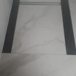 Wykończona podłoga w łazience z jasnych płytek imitujących marmur, obramowana ciemnymi płytkami i z odpływem liniowym.