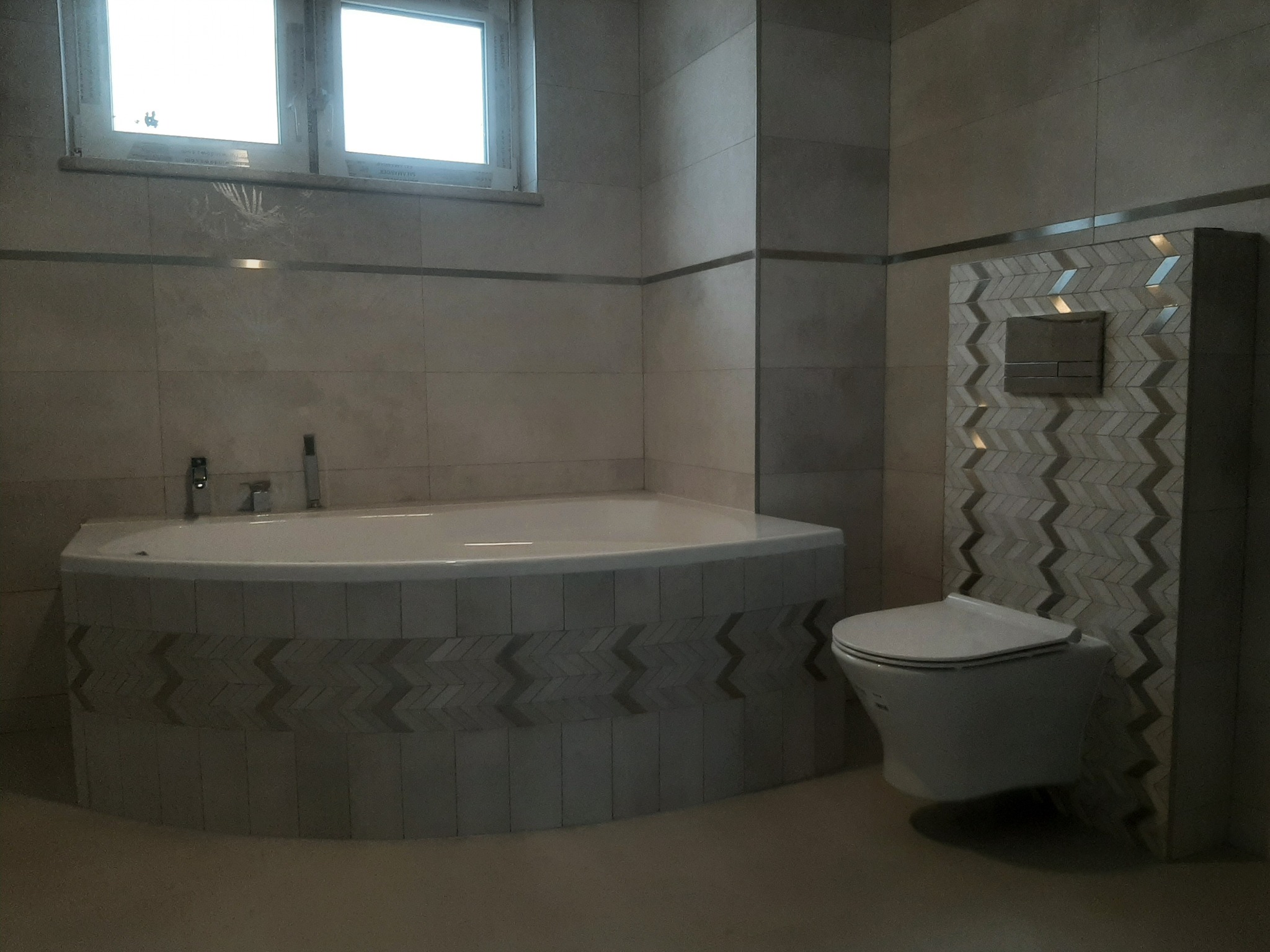 Nowoczesna łazienka z wanną narożną obudowaną płytkami z motywem chevron oraz podwieszaną toaletą z identycznym wzorem na ścianie, widoczne okno z delikatnym motywem roślinnym.