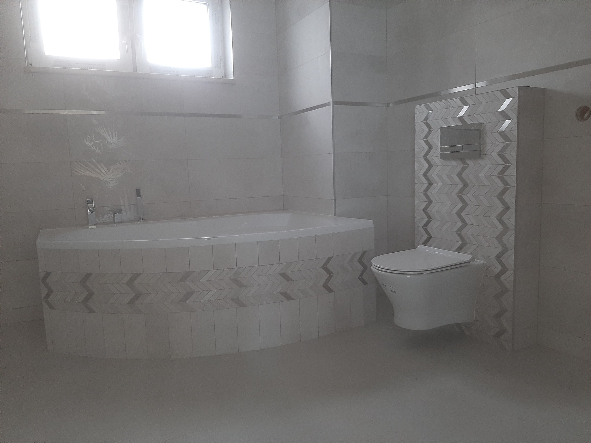 Nowoczesna łazienka w odcieniach szarości i bieli, z wanną obudowaną płytkami z motywem chevron, wiszącą toaletą i dekoracyjną ścianką z identycznym wzorem, wykończona srebrnymi listwami.