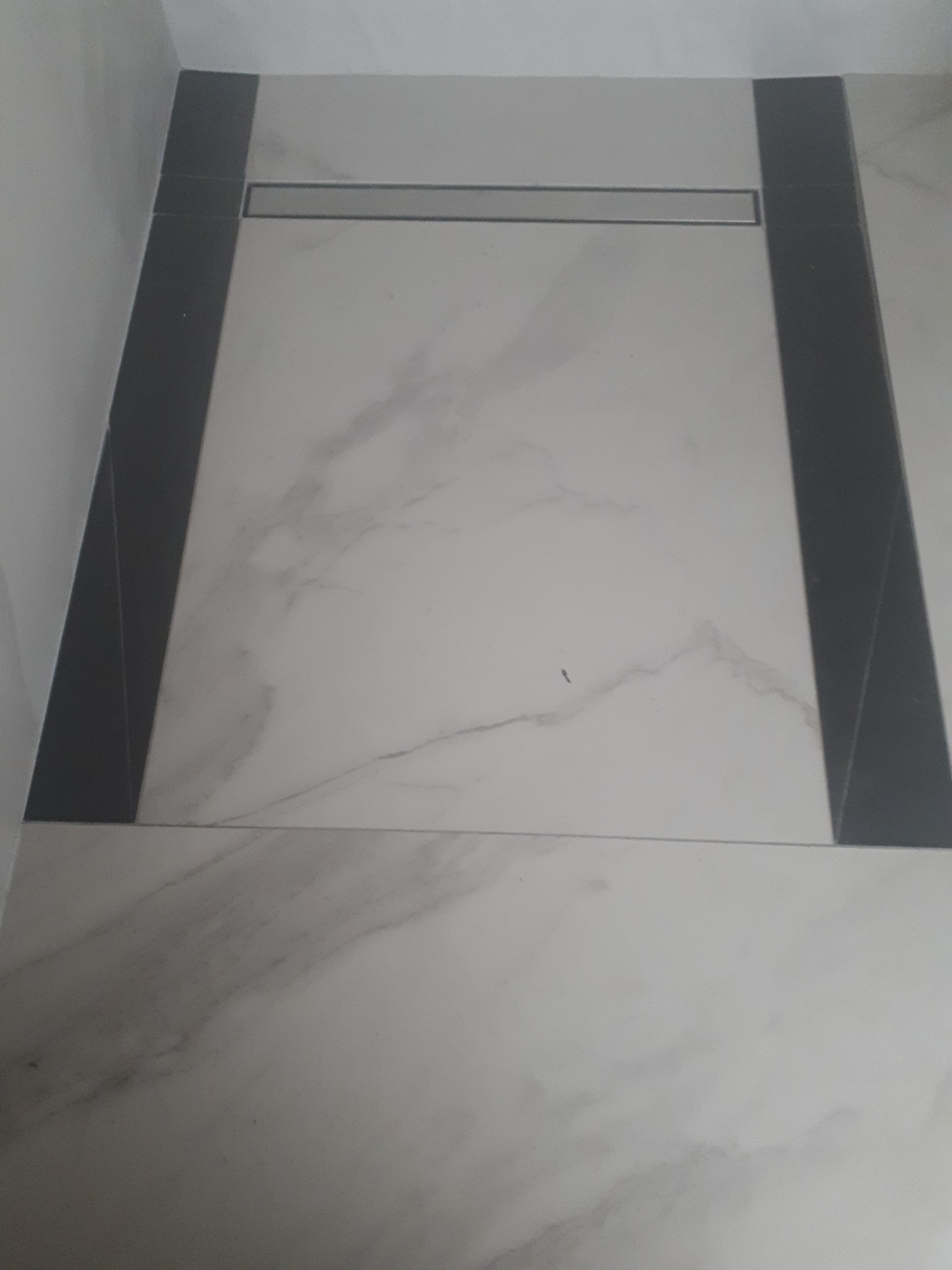 Wykończona podłoga w łazience z jasnych płytek imitujących marmur, obramowana ciemnymi płytkami i z odpływem liniowym.