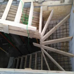 Wewnętrzna konstrukcja schodów spiralnych z drewnianych belek na etapie budowy, widok z góry na zbrojenie i formy betonowe.