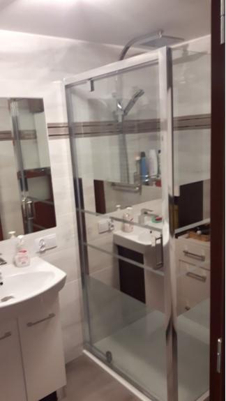 Nowoczesna kabina prysznicowa z transparentnego szkła, z widoczną deszczownicą i baterią, zamontowana w wykafelkowanej łazience z umywalką i szafkami.