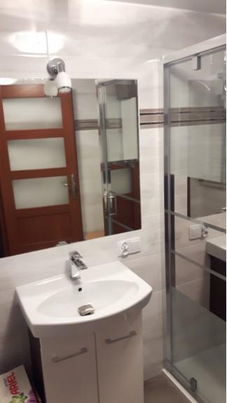 Biała umywalka z szafką w łazience, widoczne lustro odbijające drzwi i kabina prysznicowa, z pudełkiem proszku do prania Ariel u dołu.