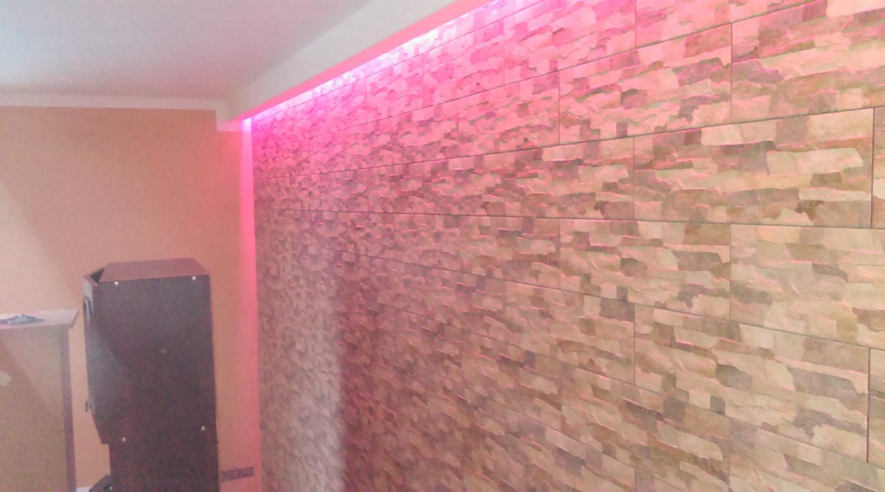 Ściana wykończona panelami imitującymi kamień, podświetlona od góry intensywnym różowym światłem LED, z widocznym fragmentem pieca lub kominka w rogu pomieszczenia.