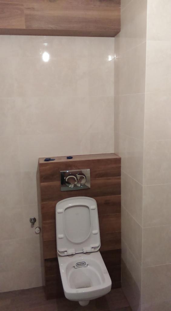 Wisząca toaleta z drewnianą obudową spłuczki i chromowanym przyciskiem, na tle beżowych, błyszczących płytek ściennych.