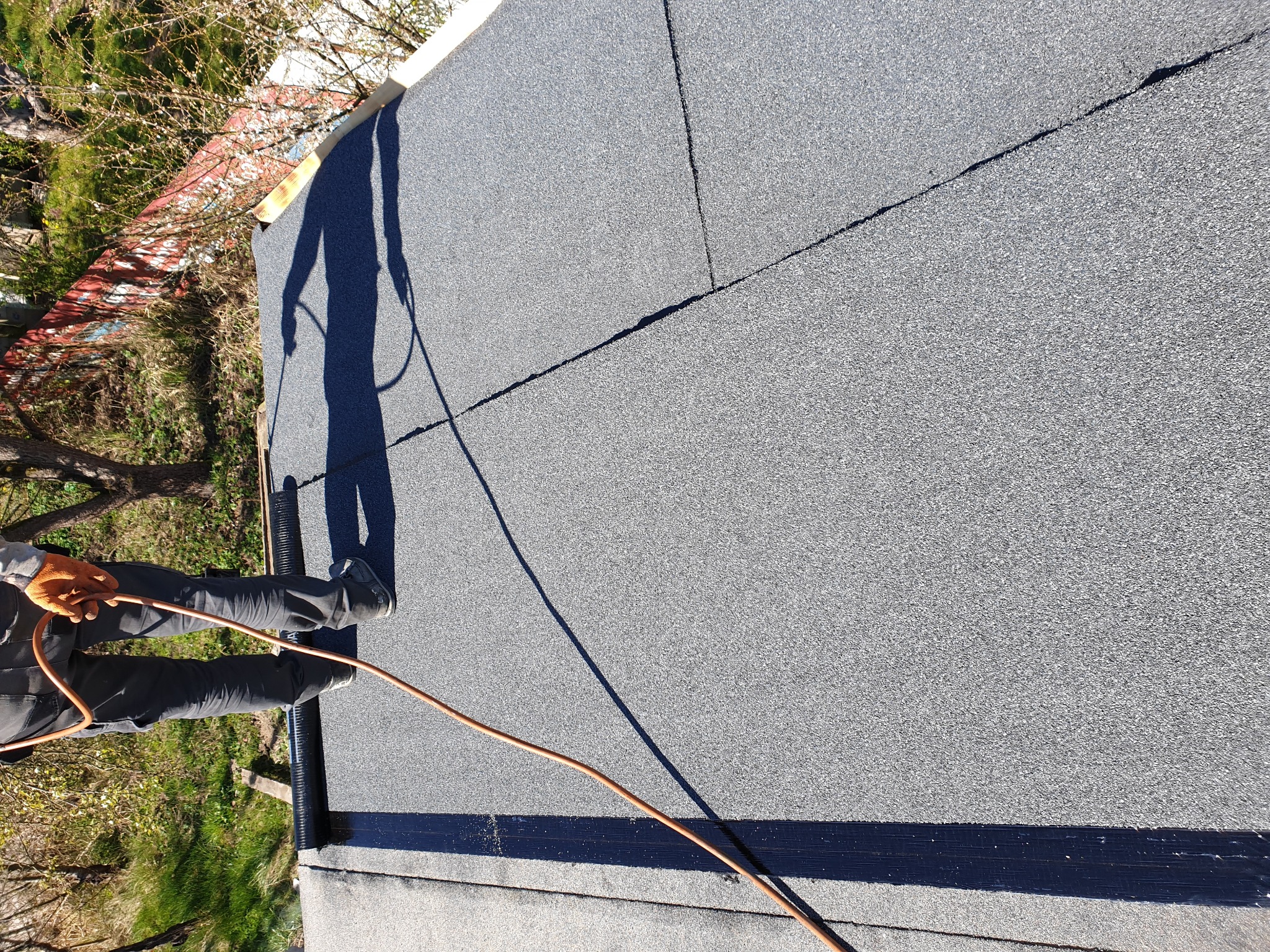 Dekarz w pomarańczowych rękawicach i ciemnych spodniach podczas prac wykończeniowych na dachu pokrytym papą, widoczny cień sylwetki i wąż do palnika.