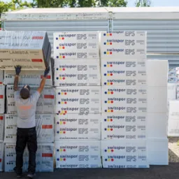 Pracownik w szarym t-shircie i czarnych spodniach układa białe, opakowane paczki materiału izolacyjnego Swissopor na palecie przed magazynem.