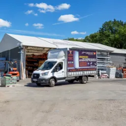 Biała ciężarówka z reklamą materiałów budowlanych na tle magazynu ze składu budowlanego, w tle widoczny wózek widłowy i palety z materiałami budowlanymi.