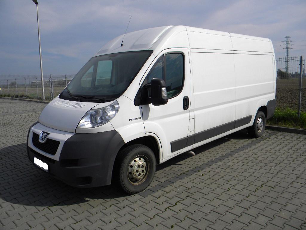 Biały samochód dostawczy Peugeot Boxer z wysokim dachem, zaparkowany na szarej kostce brukowej w pochmurny dzień, widoczne ogrodzenie z siatki i słup energetyczny w tle.