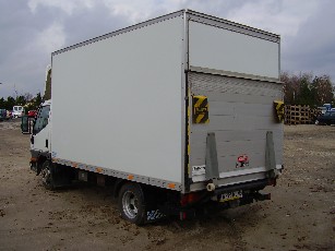 Biały samochód dostawczy z podniesioną tylną klapą, zaparkowany na żwirowym placu w pochmurny dzień. Widoczne elementy ostrzegawcze na klapie i tablica rejestracyjna.