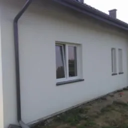 Wykończona elewacja domu jednorodzinnego z szarymi rynnami i białymi oknami, widoczne fragmenty trawnika i rury spustowej.