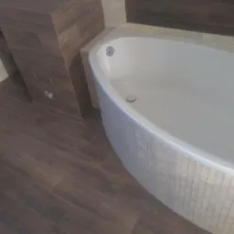 Nowoczesna biała wanna obudowana mozaiką, z brązowymi płytkami imitującymi drewno na podłodze i ścianach, widoczny fragment wnęki w ścianie.