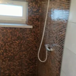 Kabina prysznicowa wyłożona mozaiką w odcieniach brązu, z widoczną słuchawką prysznicową i odpływem liniowym w podłodze.