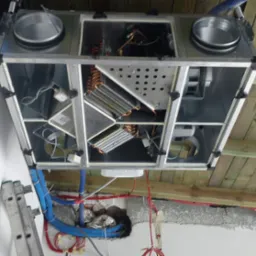 Centrala wentylacyjna podwieszona do drewnianego stropu, widoczne przewody i rury wentylacyjne, drabina oparta o ścianę, stan surowy.