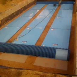 Niebieskie panele w wykopie z rurami drenażowymi, widoczne przewody elektryczne i elementy montażowe, teren budowy.