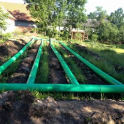 Zielone rury ułożone w ziemi na trawiastym terenie, widoczne budynki w tle, częściowo zakopane, instalacja w trakcie realizacji.