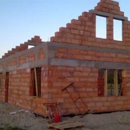 Surowy stan domu z czerwonej cegły, widoczne otwory okienne, częściowo otynkowany wieniec, teren budowy z piaskiem, w tle inne budynki i drzewa.