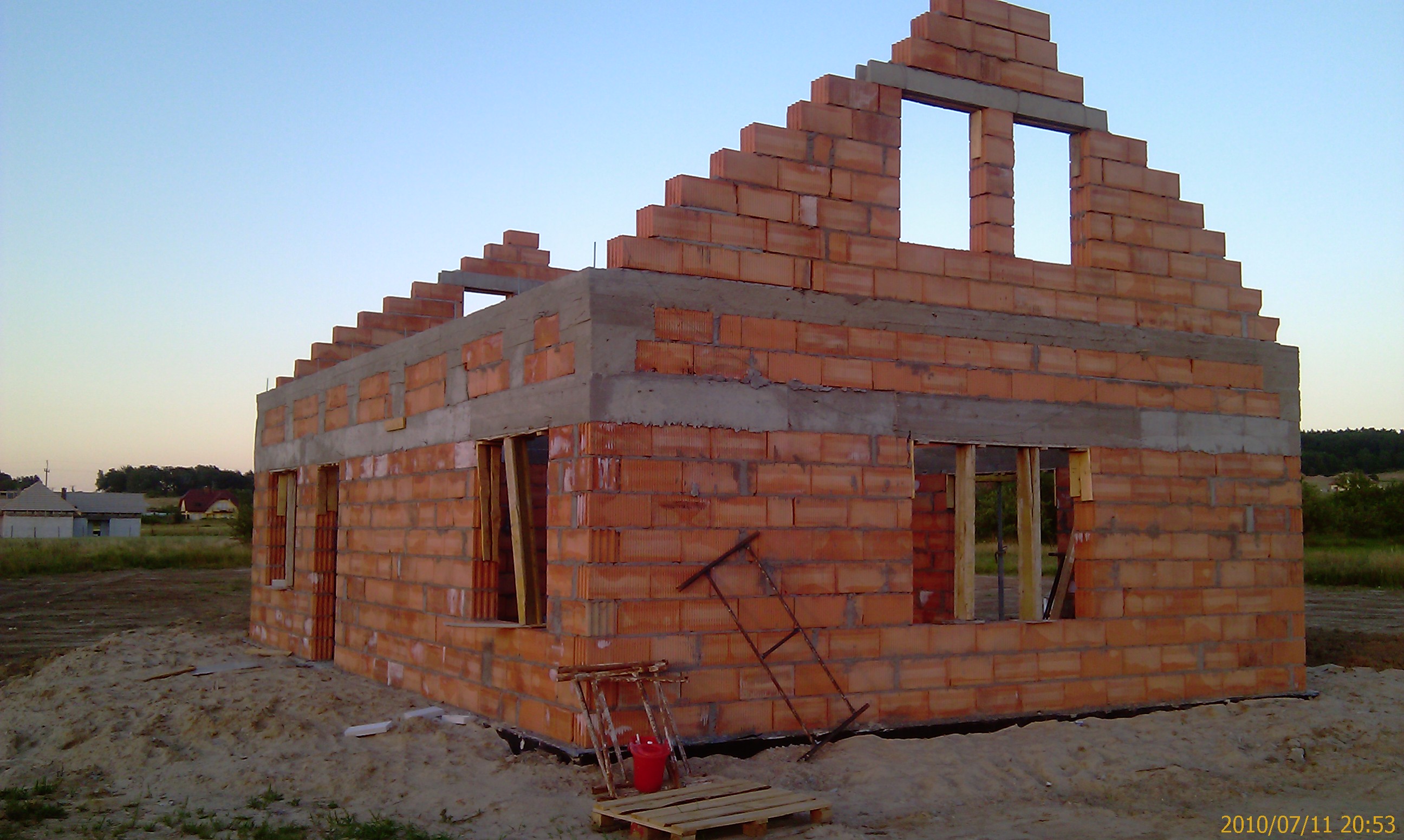 Surowy stan domu z czerwonej cegły, widoczne otwory okienne, częściowo otynkowany wieniec, teren budowy z piaskiem, w tle inne budynki i drzewa.