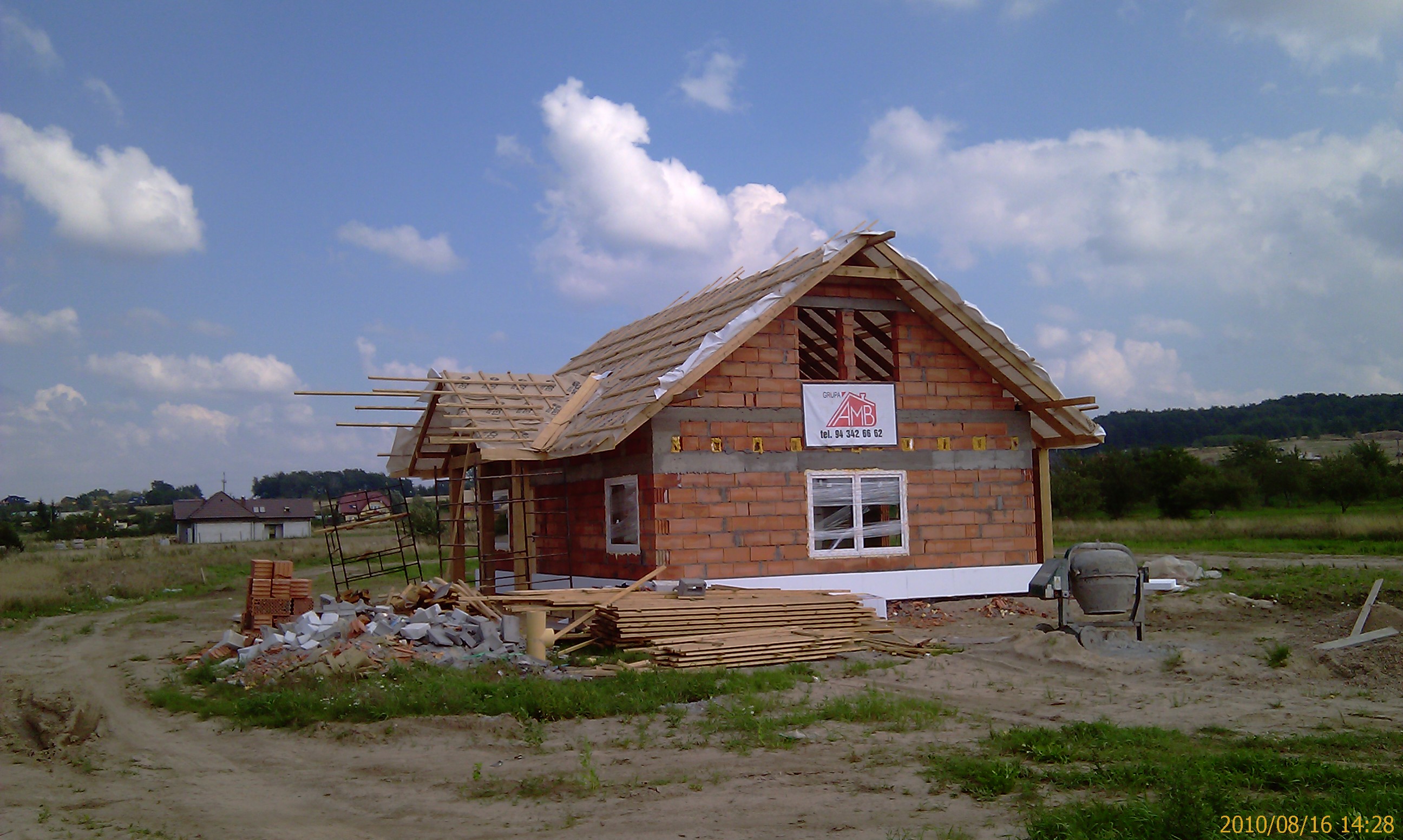 Surowy stan domu z czerwonej cegły z widocznym szkieletem dachu, drewnianymi belkami i tablicą reklamową na ścianie, w tle krajobraz z zielenią i niebem z chmurami.