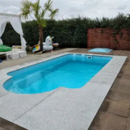 Błękitny basen ogrodowy z szarym, kamiennym obramowaniem, otoczony brązową kostką brukową, z dmuchanym jednorożcem i palmą w tle.