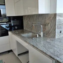 Jasnoszary blat z kamienia naturalnego z wbudowanym zlewozmywakiem i chromowaną baterią, dopasowany do ściany nad blatem z tego samego materiału, w nowoczesnej kuchni z białymi szafkami.