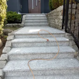 Zewnętrzne schody wejściowe do domu wyłożone szarym, nakrapianym granitem, z widocznym zwiniętym wężem ogrodowym na stopniach.