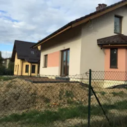 Dwa domy w różnej fazie budowy, z widoczną elewacją i dachem. Jeden dom z wykończoną elewacją w odcieniach beżu i różu, drugi w kolorze żółtym, ogrodzone siatką.