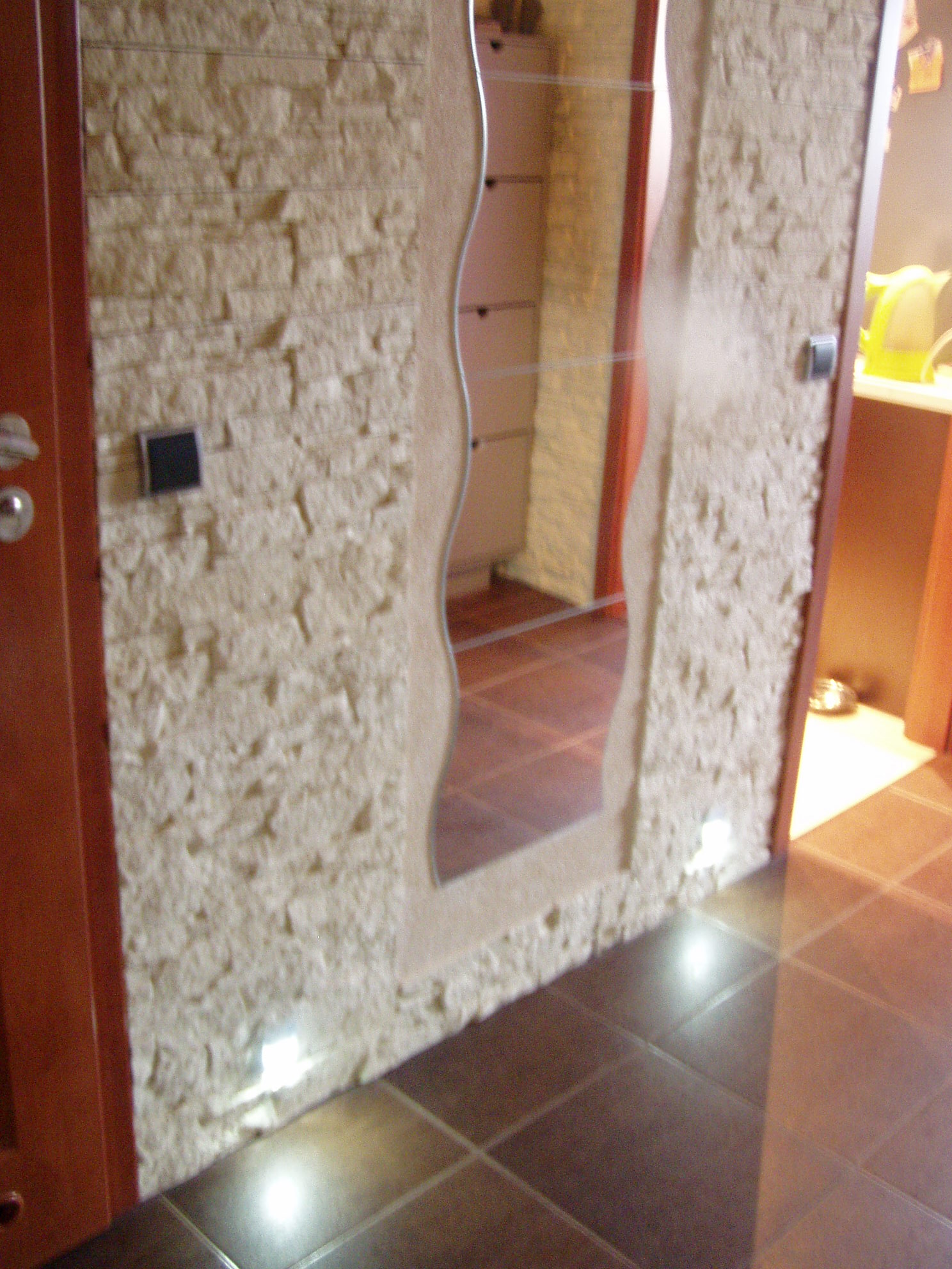 Wykończona ściana z dekoracyjną, jasną okładziną imitującą kamień, oświetlona punktowymi lampkami podłogowymi i z lustrem o falistym kształcie, brązowa podłoga z płytek.