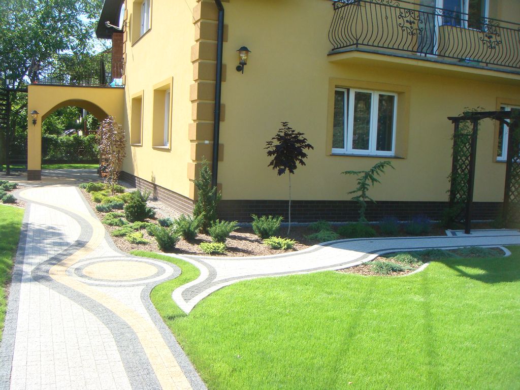 Starannie zaprojektowany ogród przydomowy z krętym chodnikiem z kostki brukowej w odcieniach szarości i beżu, prowadzącym przez rabaty z iglakami i niskimi krzewami, oraz równo przystrzyżonym...