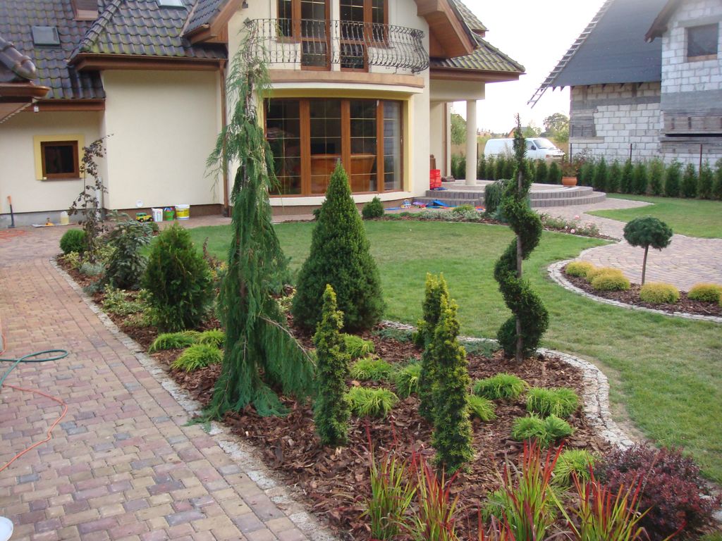 Starannie zaprojektowany ogród przydomowy z różnorodnymi roślinami iglastymi, krzewami i trawami ozdobnymi, wyściółkowany korą, z brukowanym chodnikiem i ścieżką w tle, prowadzącą do domu z balkonem.