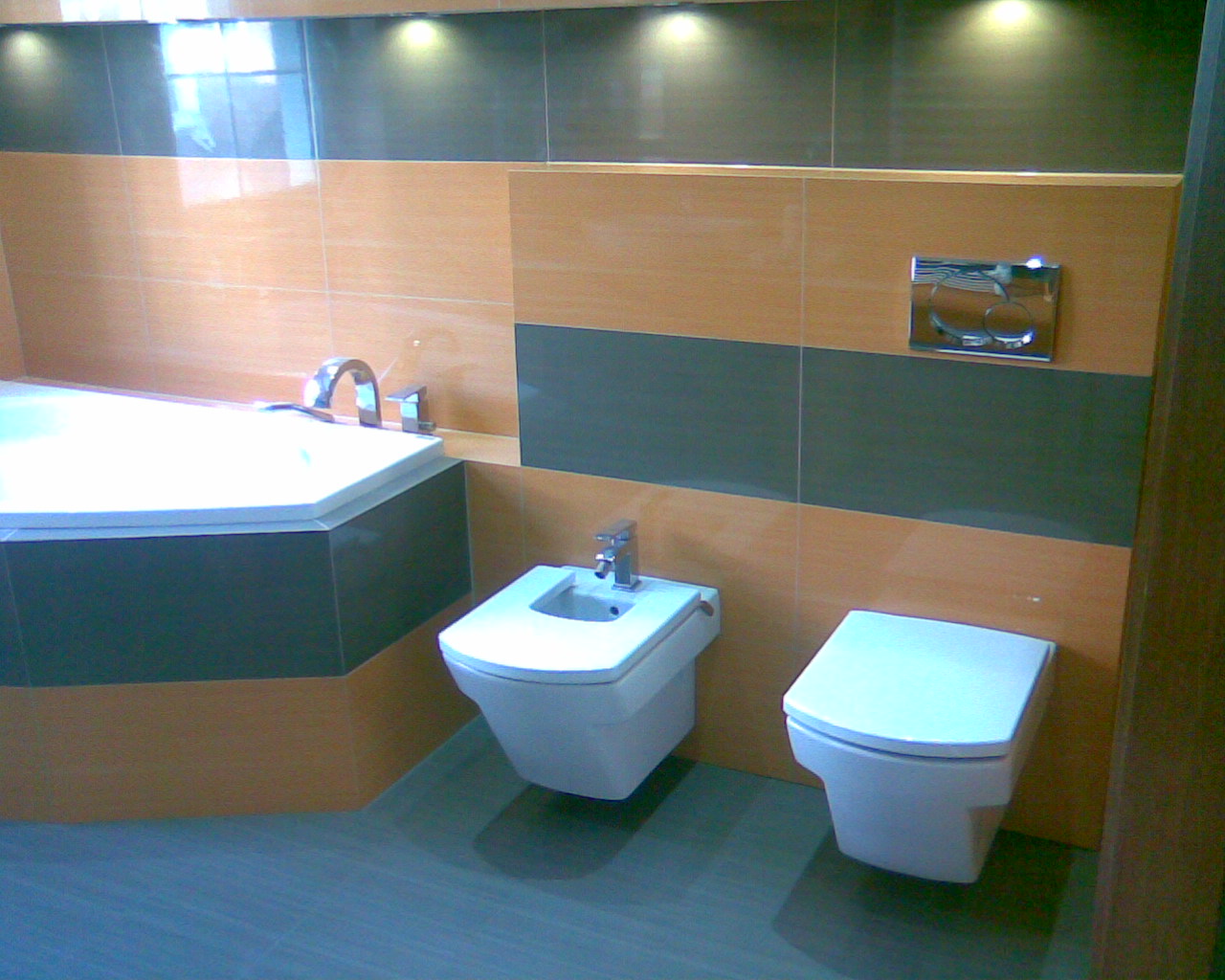 Nowoczesna łazienka z wanną narożną, bidetem i podwieszaną toaletą, wykończona płytkami w odcieniach beżu i szarości, oświetlona punktowymi lampami sufitowymi.