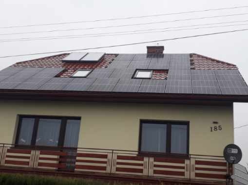 Dom jednorodzinny z częściowo zainstalowanymi panelami słonecznymi na dachu z czerwonej dachówki, widoczne okna dachowe i numer domu '185'.